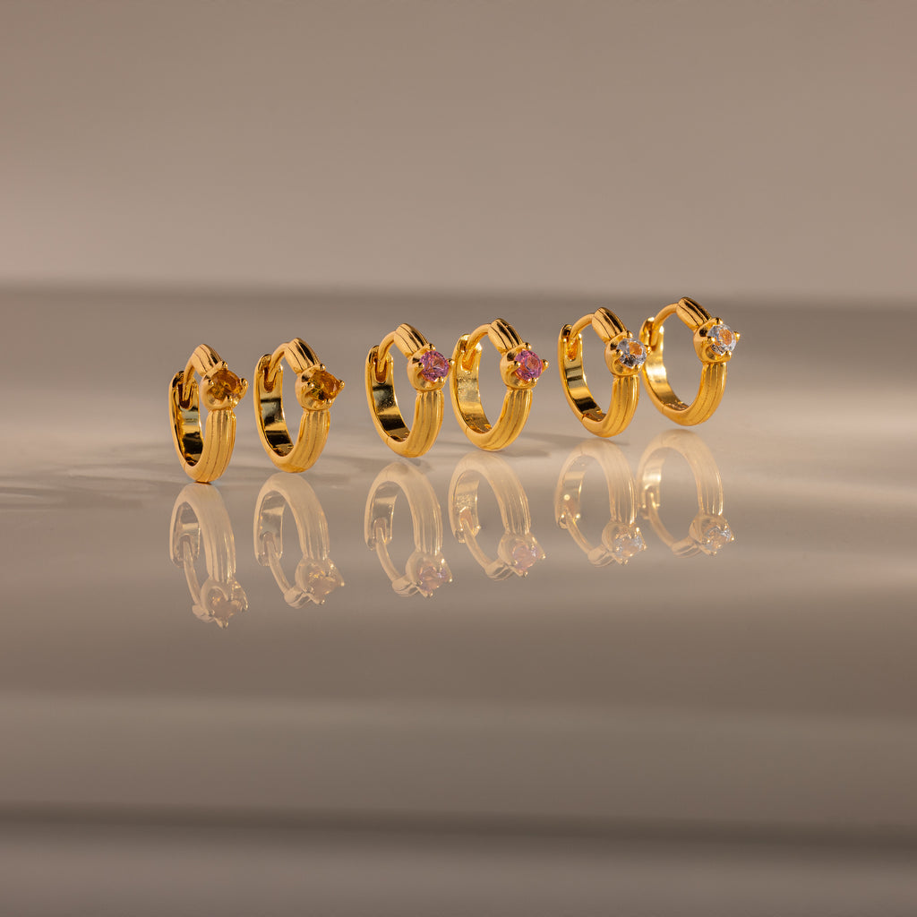 The Ribbed Ruby Huggies, inspired by birthstone jewelry, are displayed in a row with a sleek reflection on a smooth surface.