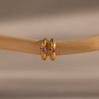 Gold hoop earrings with ruby gemstones, inspired by Ribbed Ruby Huggies, displayed on a beige branch against a neutral background—a stunning piece of birthstone jewelry.