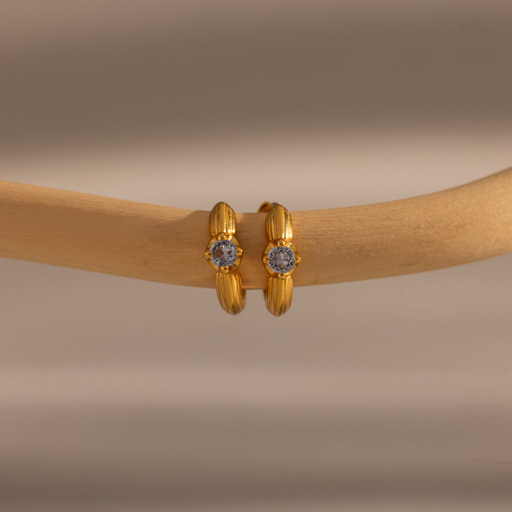 Close-up of two Ribbed Sapphire Huggies gold hoop earrings, each adorned with a small sapphire gemstone and displayed on a beige wooden rod. Perfect for lovers of personalized jewelry.