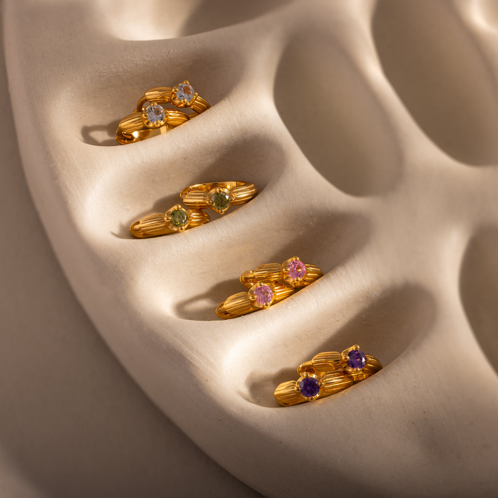 Five gold rings with colorful gemstones on a beige, textured surface—perfect as a meaningful gift or paired with Ribbed Amethyst Huggies and gold hoop earrings.