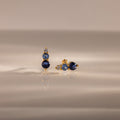 Ombre Blue Zircon Flatback Studs feature three blue zircon gems and a small clear stone on a gold setting—an elegant, personalized gift idea, beautifully displayed on a reflective surface.