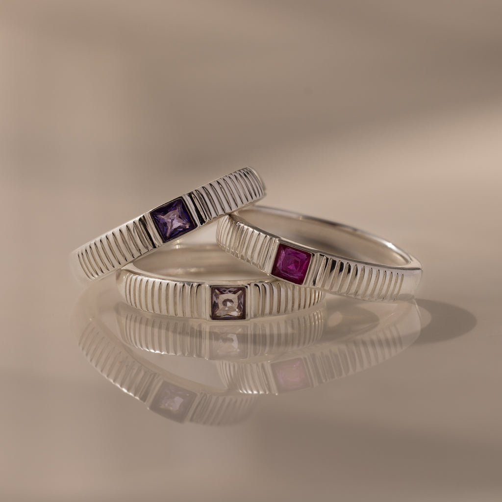 The Ribbed Amethyst Band is a stylish silver stacking ring featuring a square purple gemstone, perfect for adding birthstone elegance to any look.