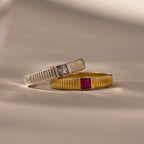 The Ribbed Ruby Band—a gold ring with a ribbed band and red gem—is shown alongside a silver birthstone ring with a ribbed band and clear square cut stone, both displayed on a beige surface.
