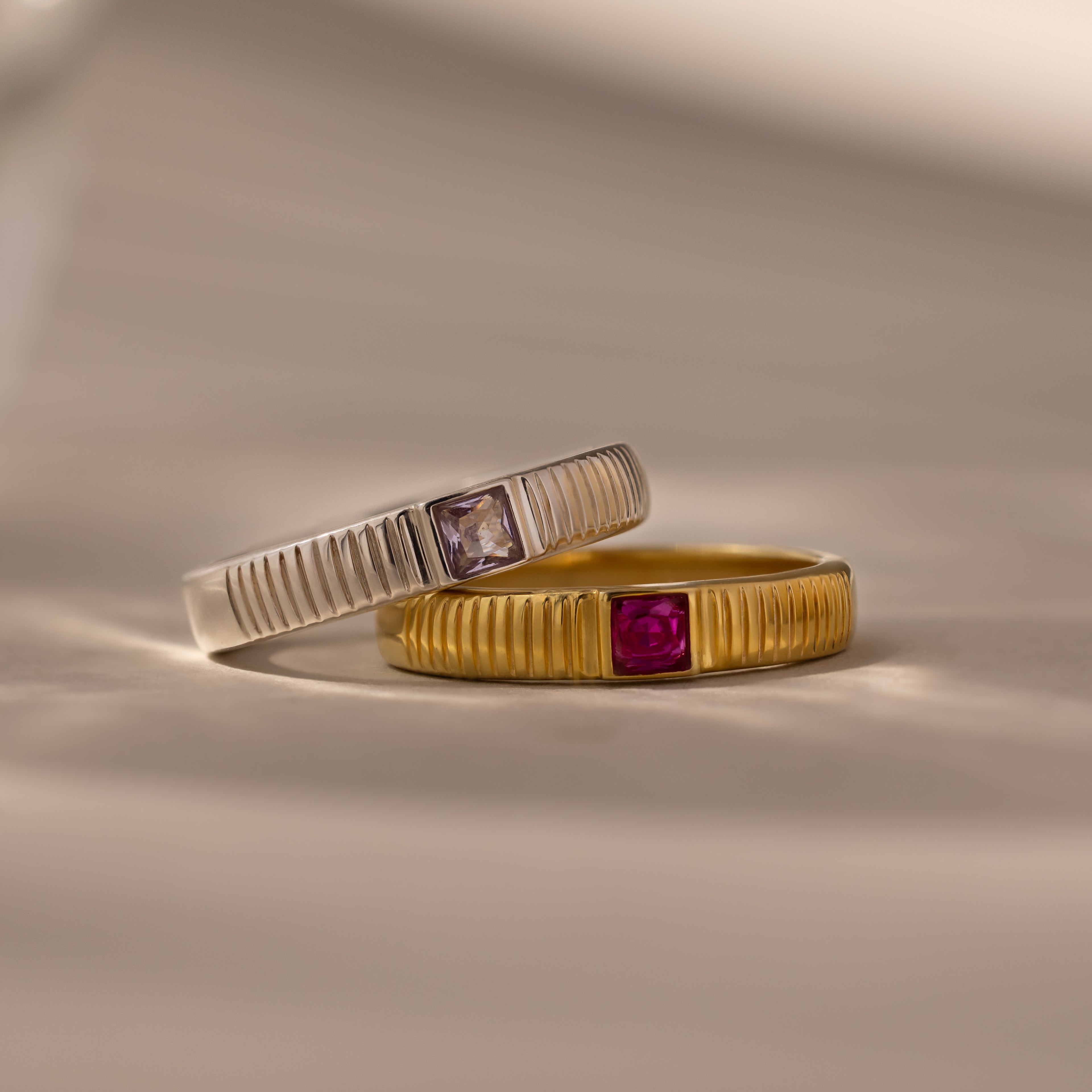The Ribbed Ruby Band—a gold ring with a ribbed band and red gem—is shown alongside a silver birthstone ring with a ribbed band and clear square cut stone, both displayed on a beige surface.
