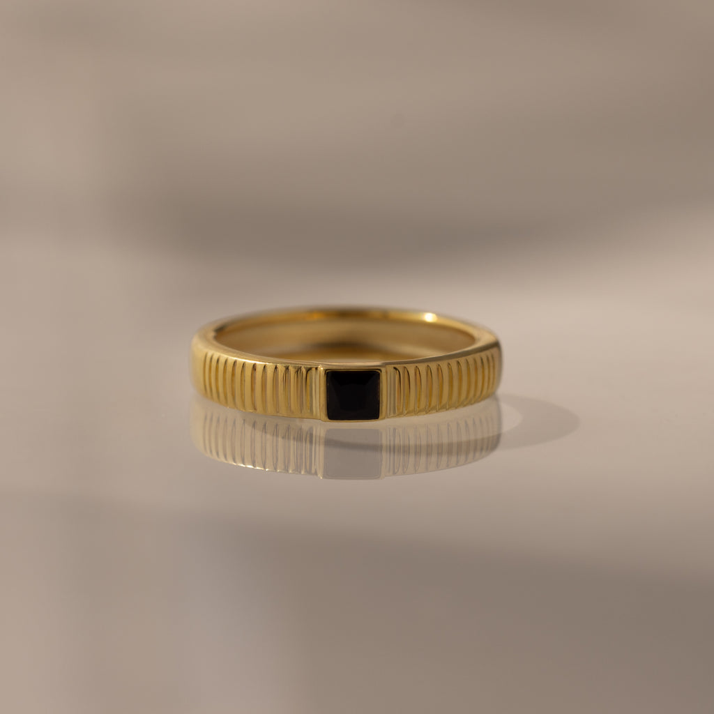 The Ribbed Onyx Band is a gold birthstone ring featuring a black square-cut onyx set in the center, beautifully displayed on a smooth, reflective surface.