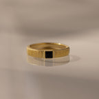 The Ribbed Onyx Band is a gold birthstone ring featuring a black square-cut onyx set in the center, beautifully displayed on a smooth, reflective surface.