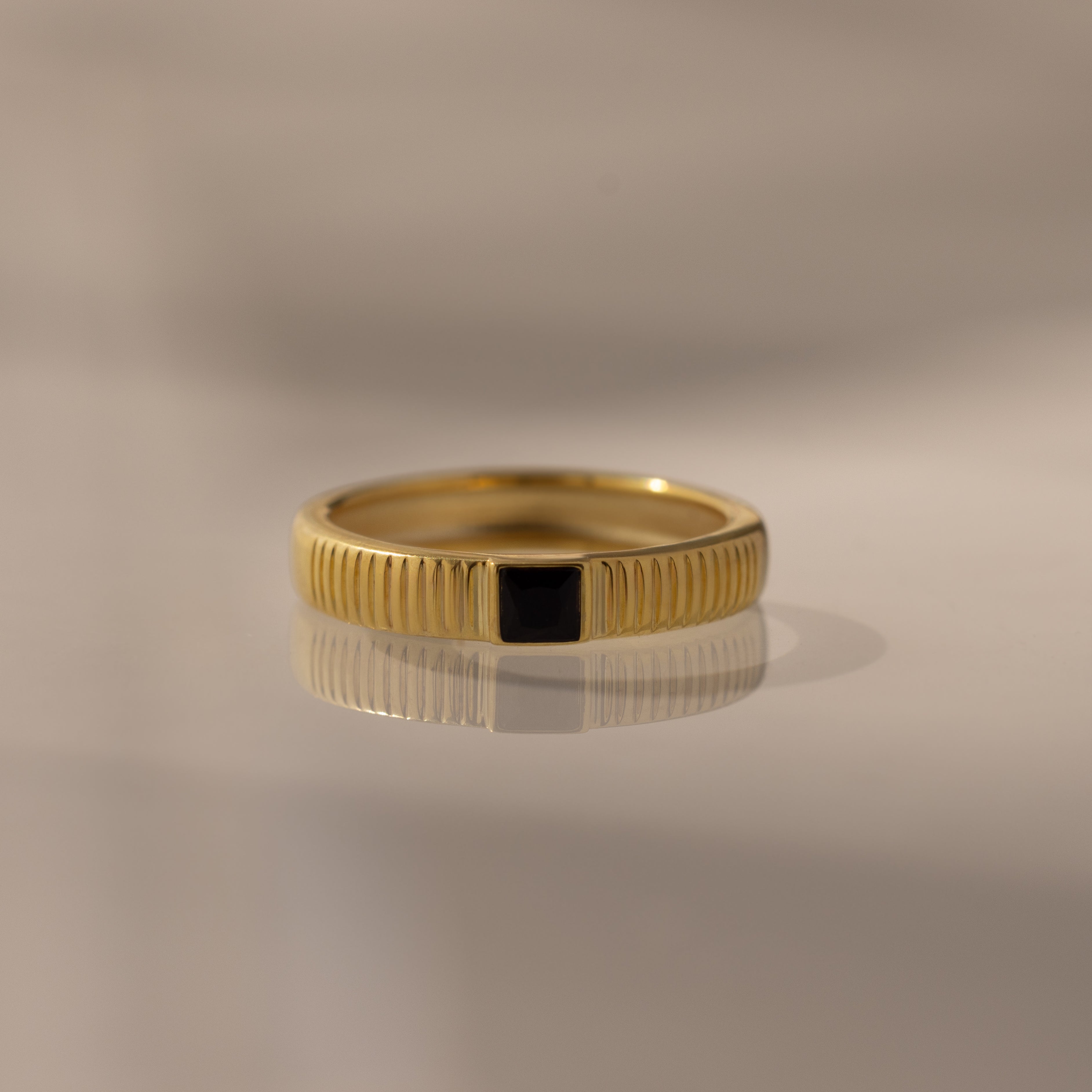 The Ribbed Onyx Band is a gold birthstone ring featuring a black square-cut onyx set in the center, beautifully displayed on a smooth, reflective surface.