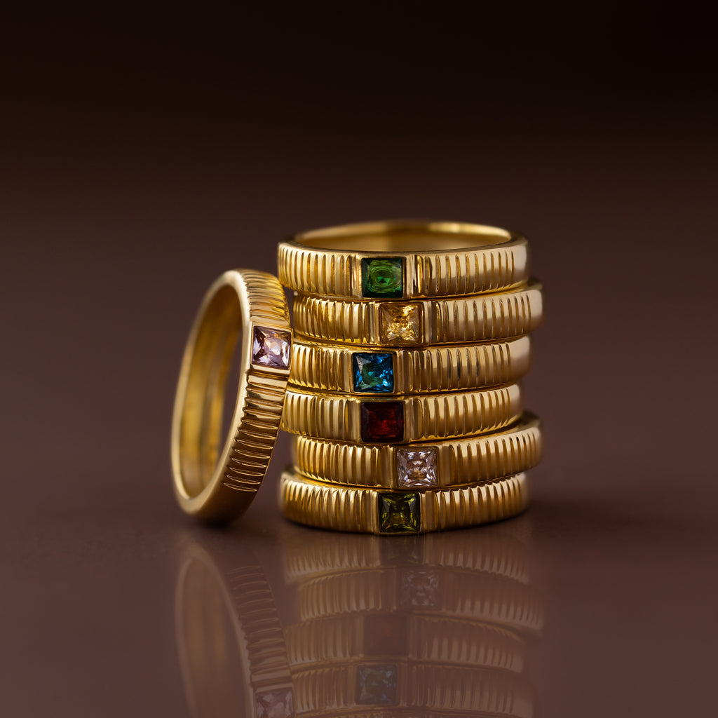 The Ribbed Amethyst Band, stacked with various colored gemstone rings, is elegantly displayed on a reflective brown surface.
