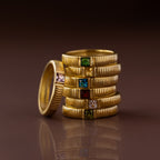 The Ribbed Blue Zircon Band, part of a gold stacking ring set with colorful square gemstones, is arranged neatly on a reflective brown surface.
