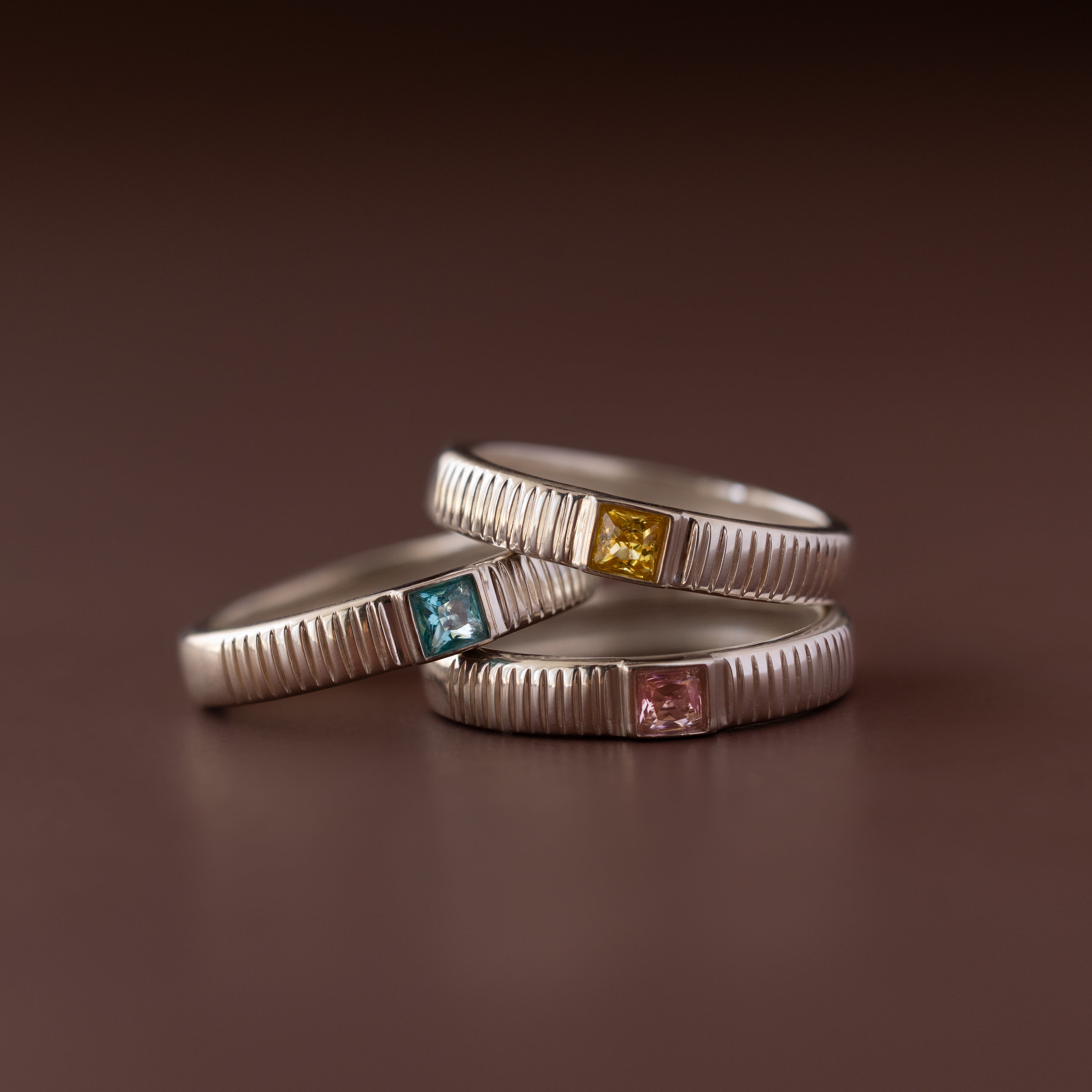 Three Ribbed Topaz Bands, each with a vibrant square-cut stone in blue, yellow, and pink, are stacked elegantly on a brown surface.