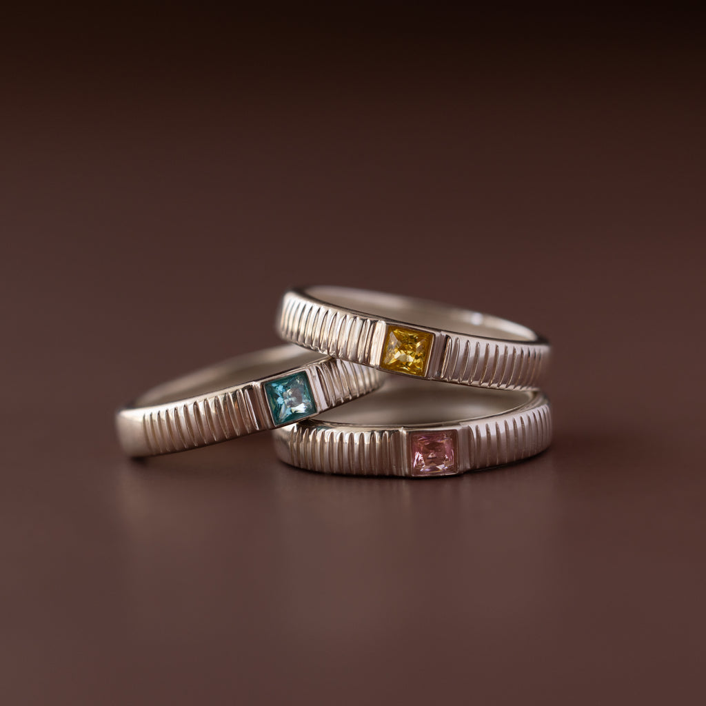 The Ribbed Birthstone Band features three silver stacking rings with textured bands, each set with a different colored square gemstone, for a unique birthstone ring style.