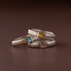 The Ribbed Birthstone Band features three silver stacking rings with textured bands, each set with a different colored square gemstone, for a unique birthstone ring style.