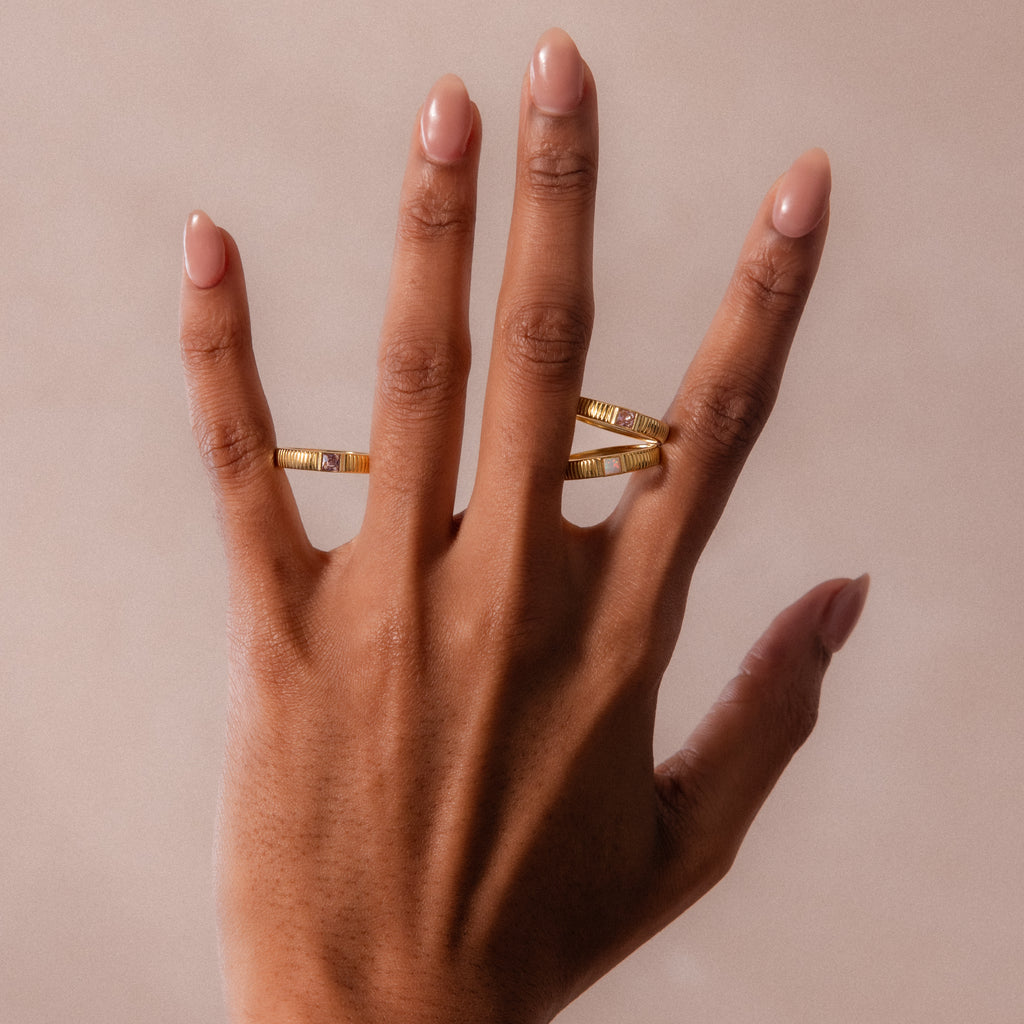 A hand with nude nails showcases stacking rings—a gold band and the Ribbed Diamond Band—on the middle and ring fingers against a neutral background.