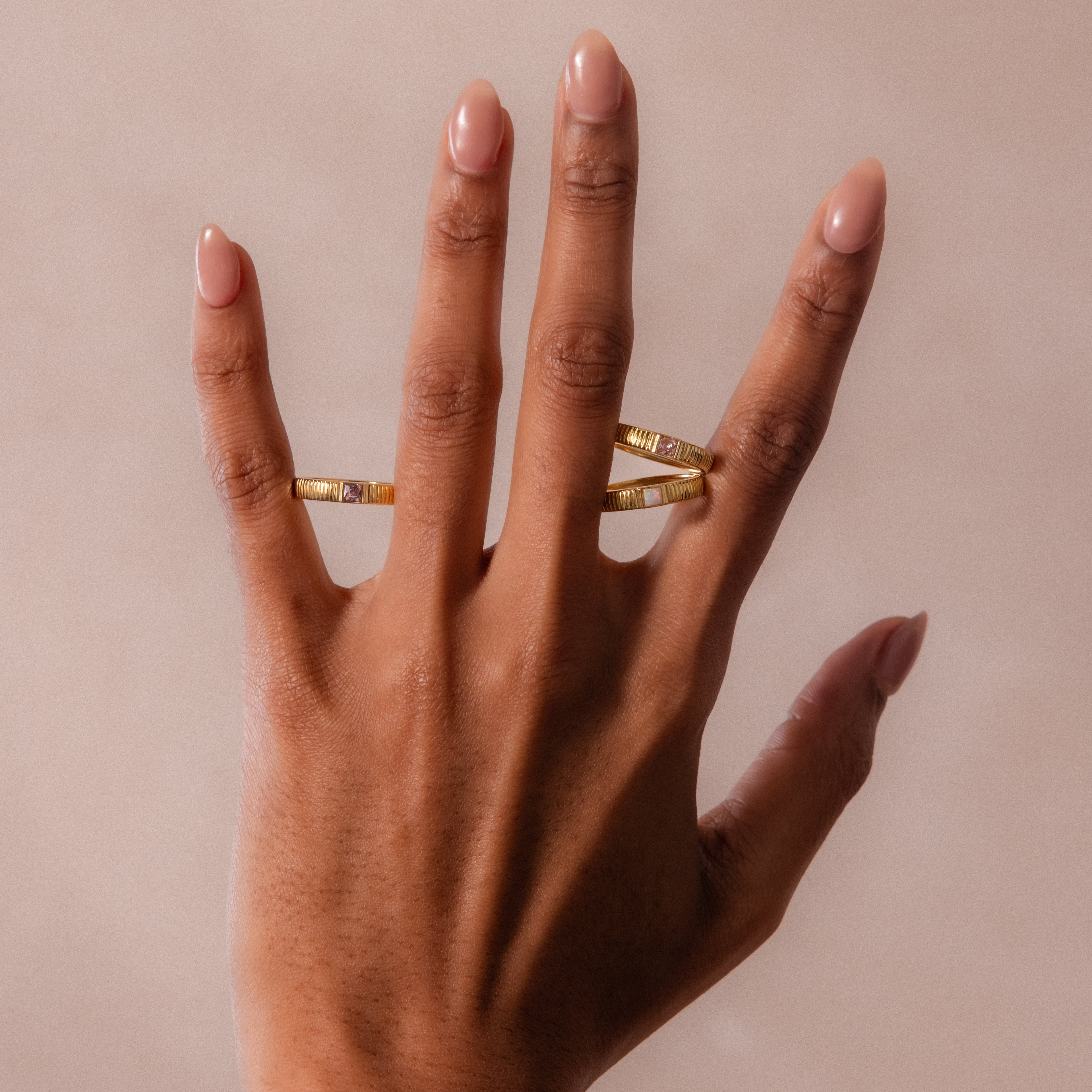 A hand with nude nail polish wears the Ribbed Opal Band and two gold stacking rings on the middle and ring fingers, set against a beige background.