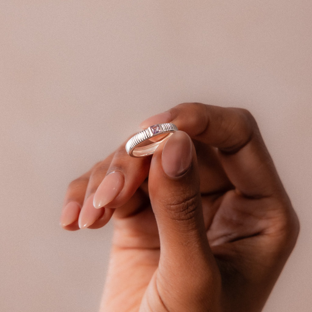 A hand with manicured nails holds the Ribbed Amethyst Band, a silver stacking ring featuring a small amethyst, against a neutral background.