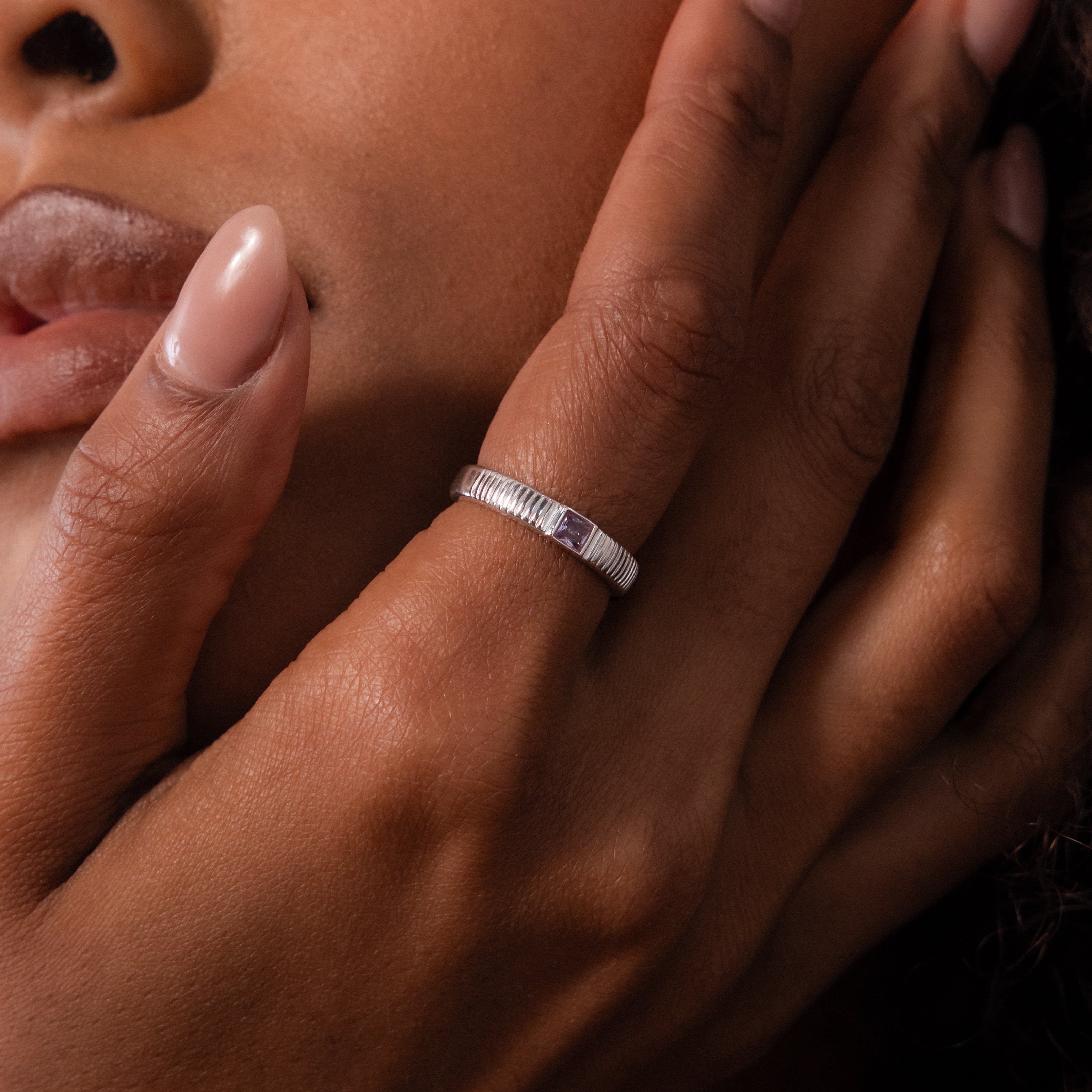 A close-up of a hand with manicured nails gently touching the face, showcasing the Ribbed Amethyst Band.