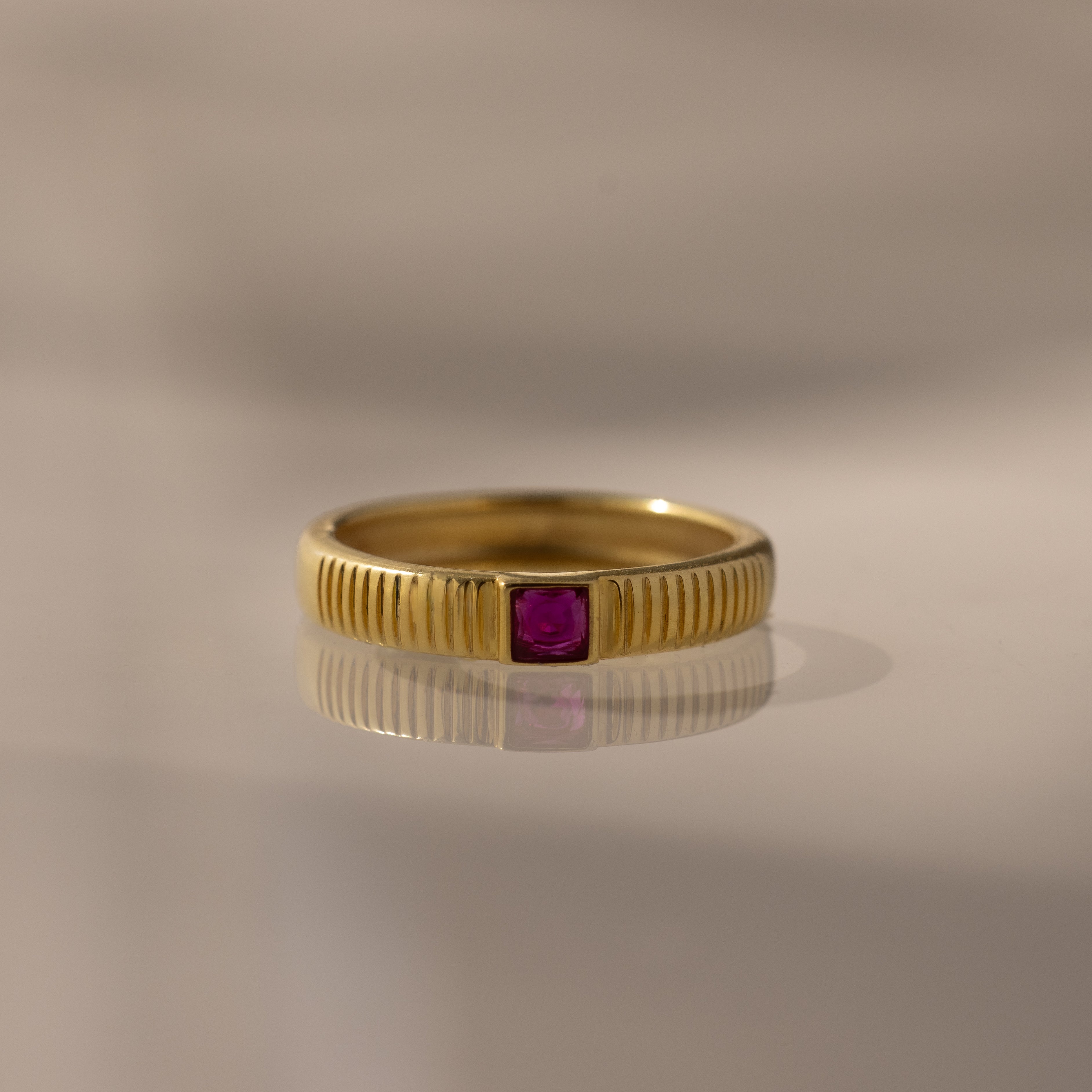 The Ribbed Ruby Band is a gold ring with a ribbed band and a small pink square-cut stone set at its center.