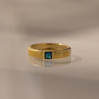 The Ribbed Blue Zircon Band, a gold stacking ring featuring a square blue gemstone at its center, is displayed on a reflective surface.