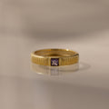 The Ribbed Amethyst Band is a gold stacking ring with vertical grooves and a small square purple amethyst in the center, shown on a reflective surface.