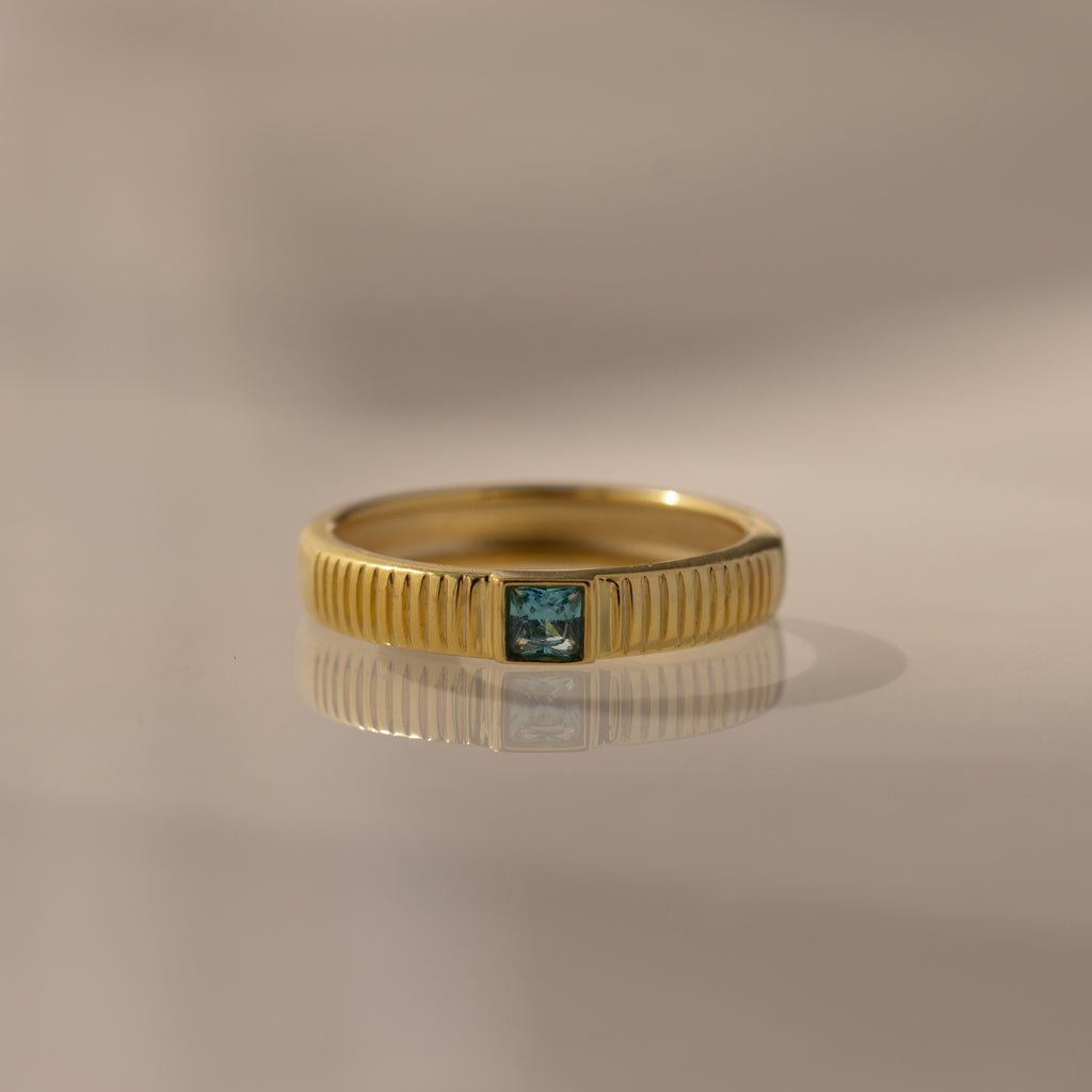 The Ribbed Aquamarine Band features vertical gold grooves and a central blue gemstone, ideal as a birthstone or stacking ring, displayed against a neutral background.