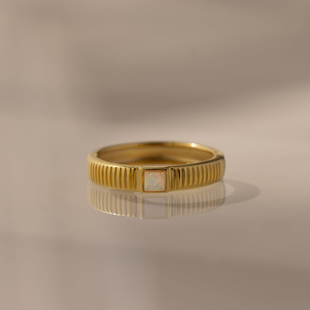 The Ribbed Opal Band features a gold ribbed band and a small rectangular opal stone, beautifully reflected on a smooth surface.