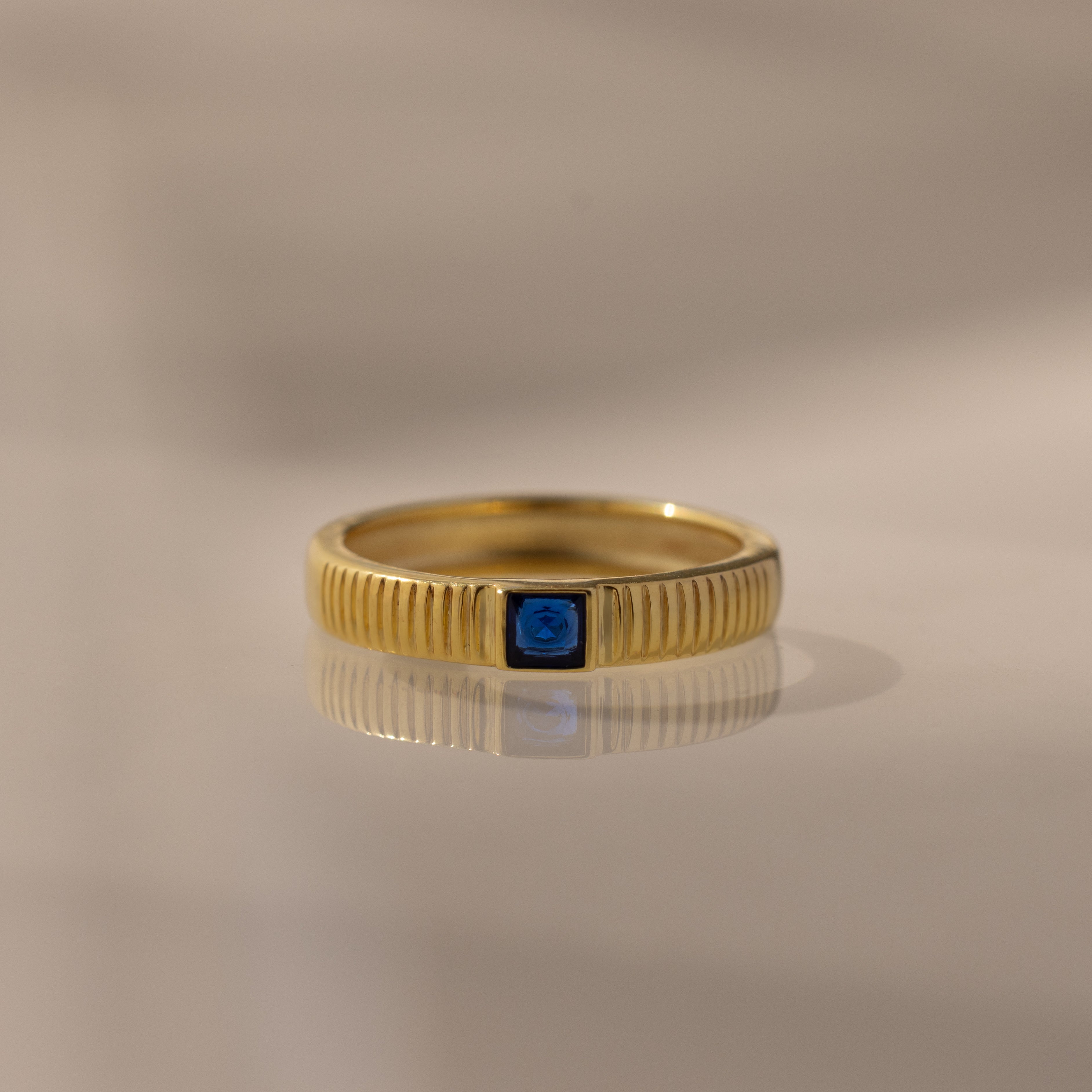 The Ribbed Sapphire Band features a gold ribbed, textured band with a small square blue sapphire at the center, ideal as a birthstone ring and shown on a neutral background.