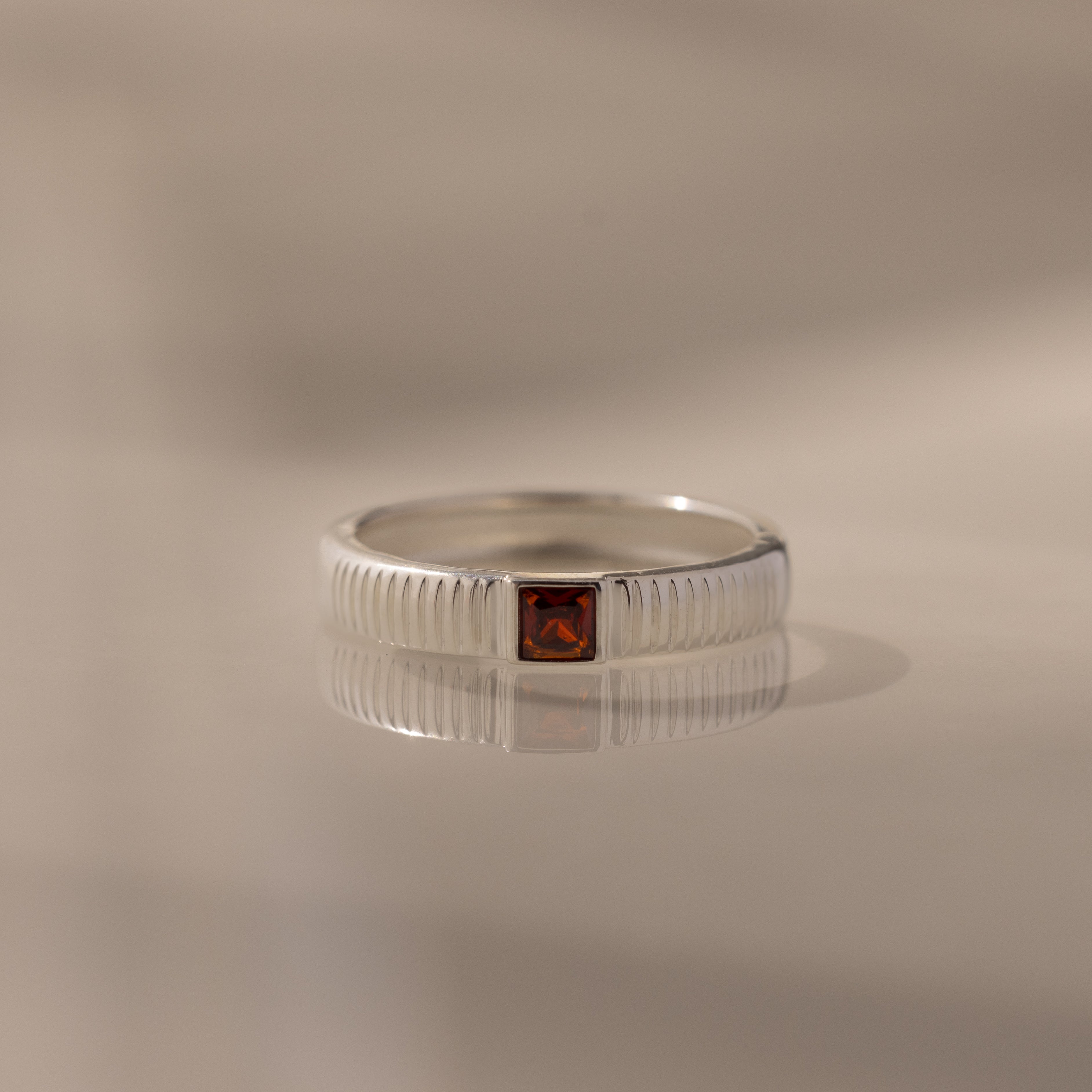 The Ribbed Garnet Band is a silver birthstone ring featuring a square red garnet gemstone in the center, displayed on a reflective surface.