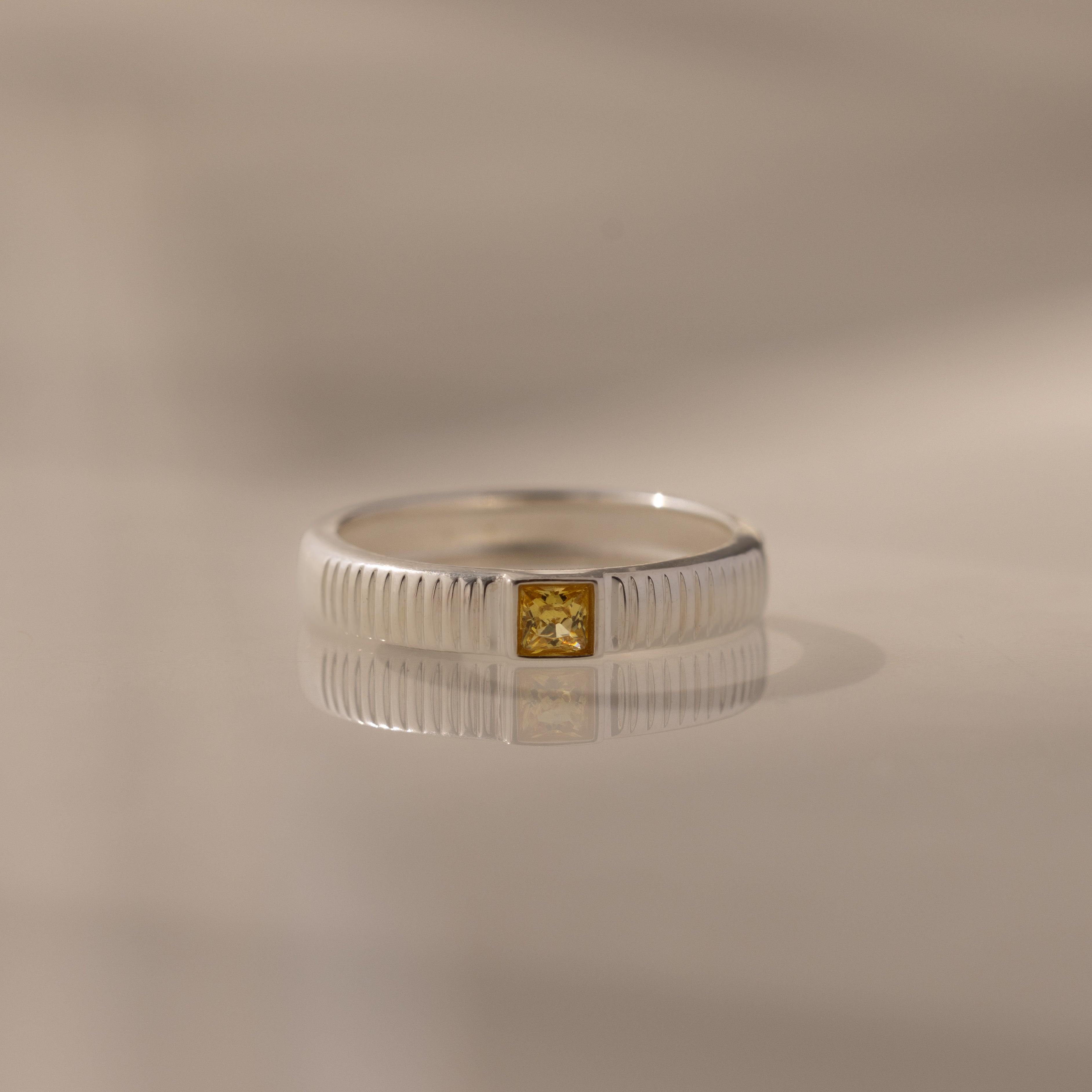 The Ribbed Topaz Band features a silver ribbed band and a square-cut yellow gemstone, displayed on a soft, neutral background with a subtle reflection.