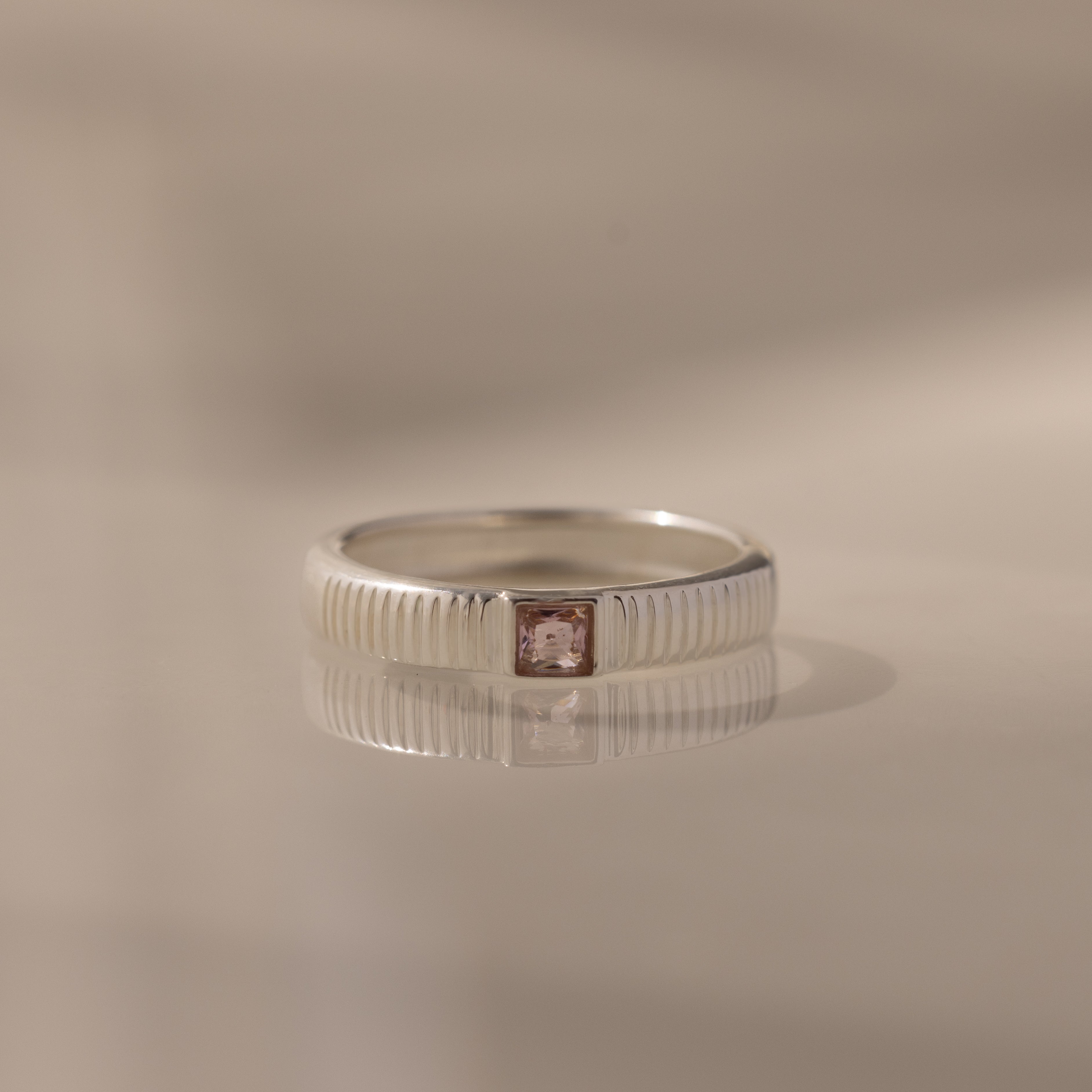 The Ribbed Pink Tourmaline Band features a silver ribbed ring with a small square pink tourmaline gemstone at the center, ideal as a birthstone ring and beautifully displayed on a reflective surface.