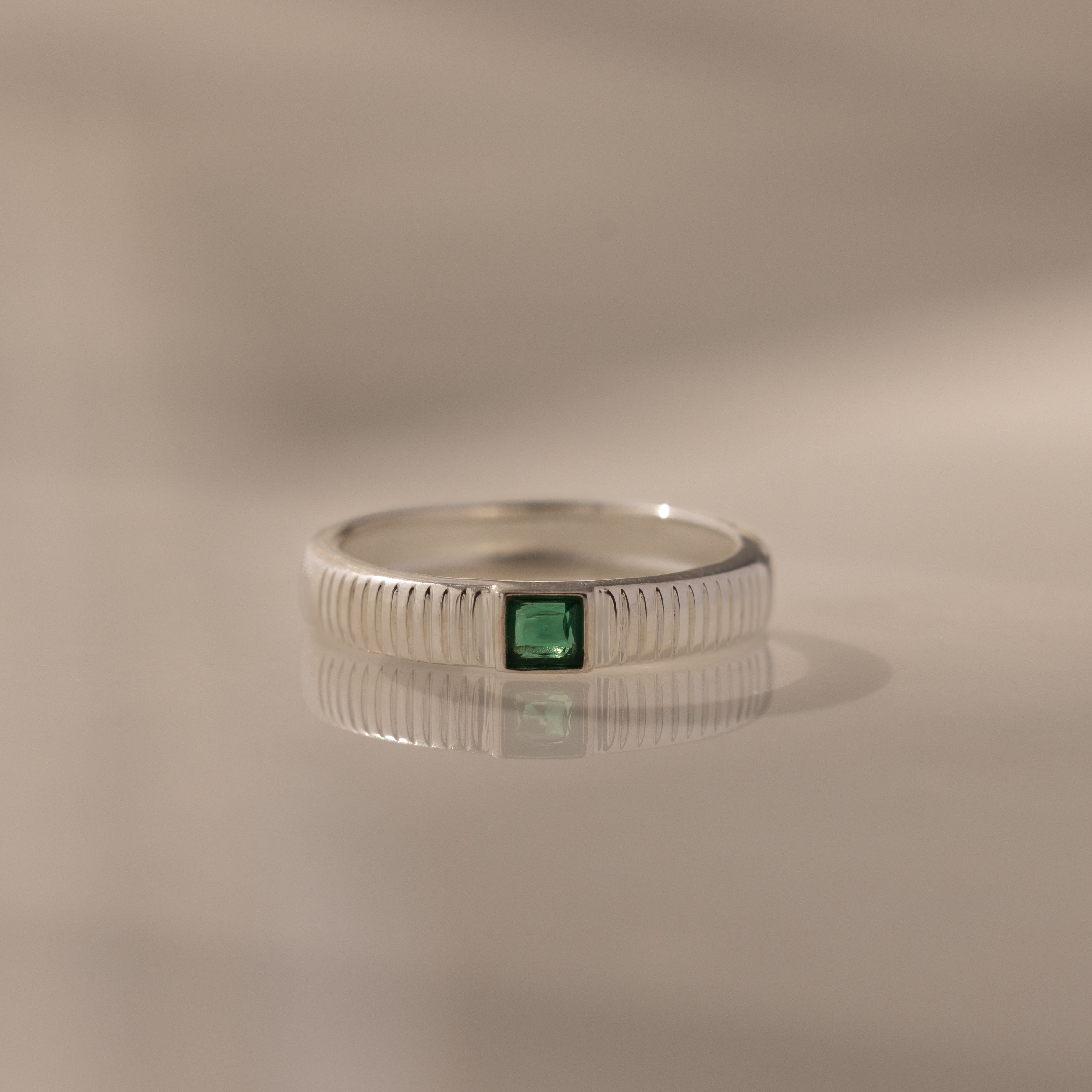 The Ribbed Emerald Band, a silver ring featuring a square-cut green gemstone, is displayed on a reflective surface under soft lighting.