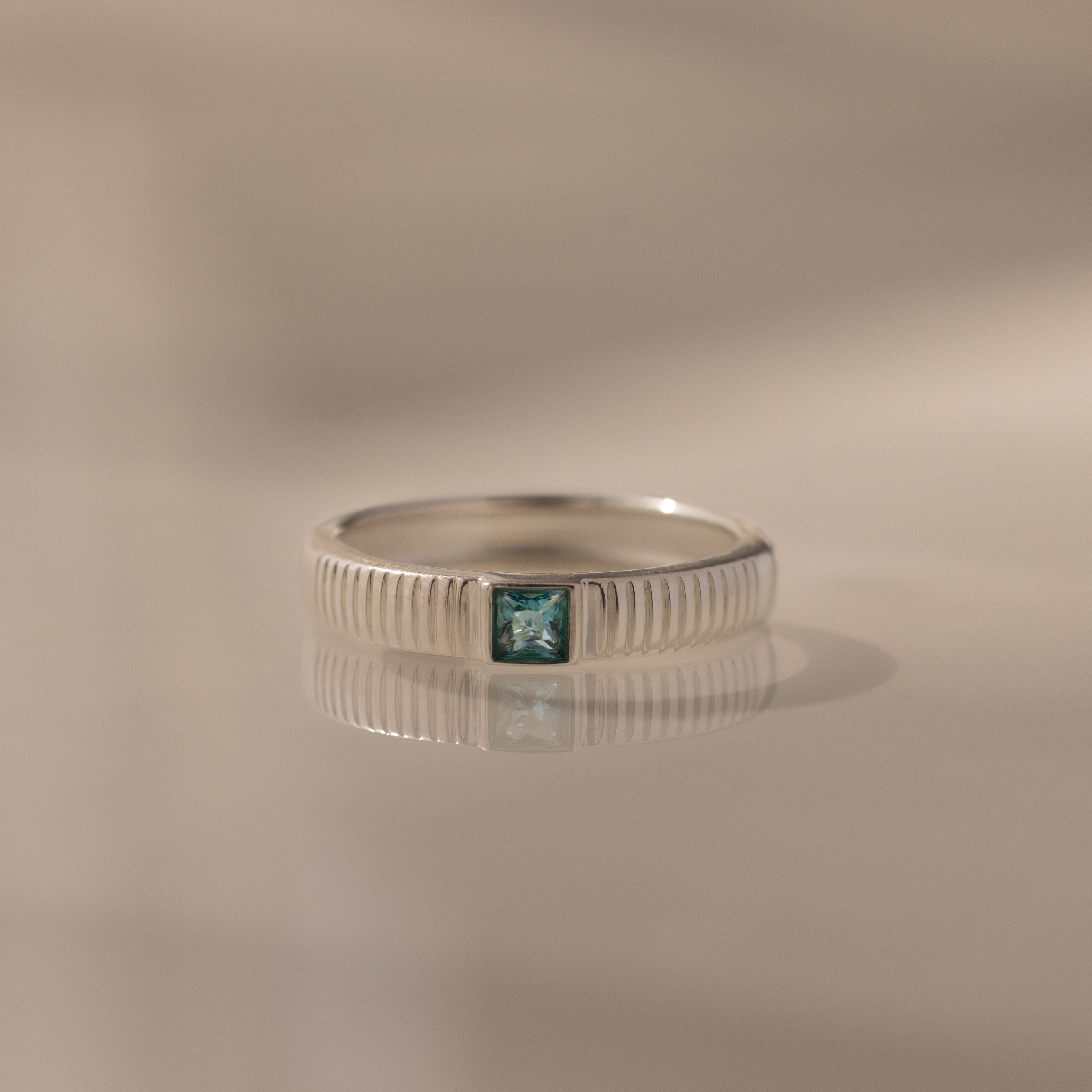 The Ribbed Aquamarine Band features a square blue aquamarine gemstone set in a silver band with ribbed detailing, beautifully displayed on a reflective surface—ideal as a modern birthstone ring.