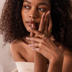 A woman with curly hair touches her face and looks to the side, wearing the Ribbed Name Signet Ring in gold against a neutral background.