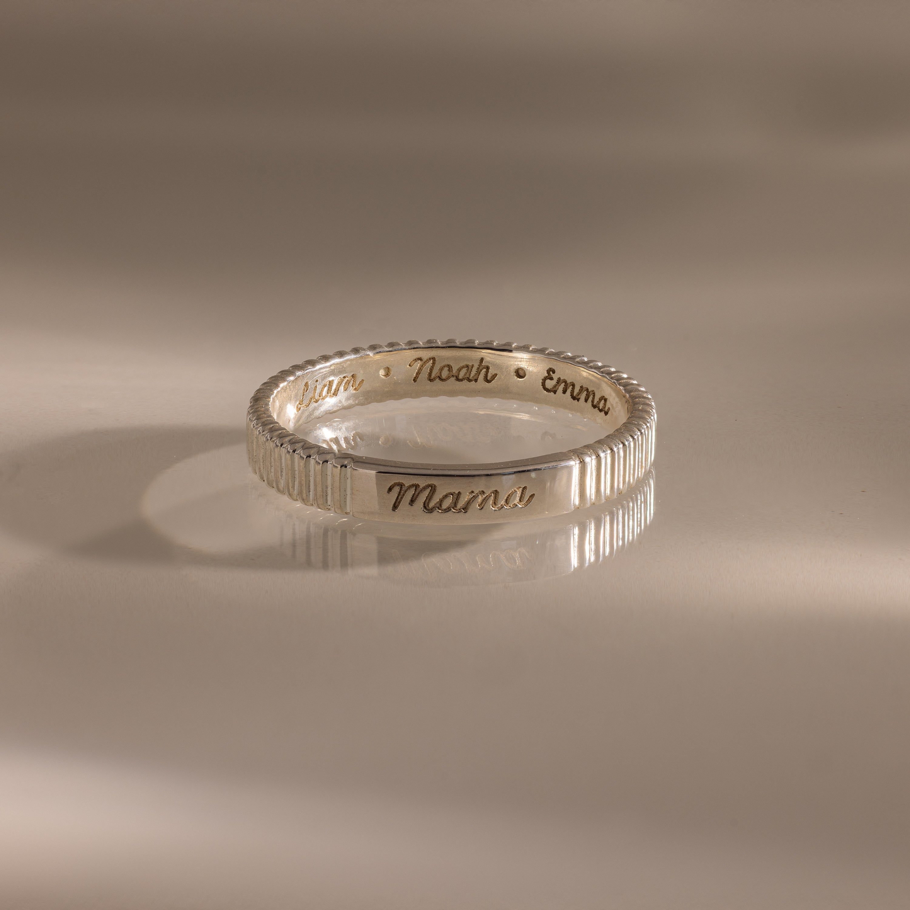 The Ribbed Name Ring in Silver, engraved with Mama, Noah, and Emma, is shown on a neutral background.