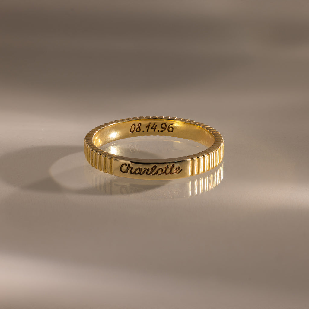 The Ribbed Name Ring in 18K Gold, engraved with "Charlotte" and the date 08.14.96, rests on a smooth, reflective surface—an elegant example of personalized jewelry.