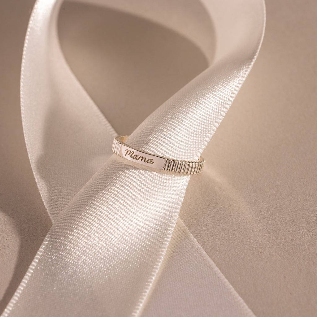 A silver Ribbed Name Ring engraved with Mama rests on a white satin ribbon.