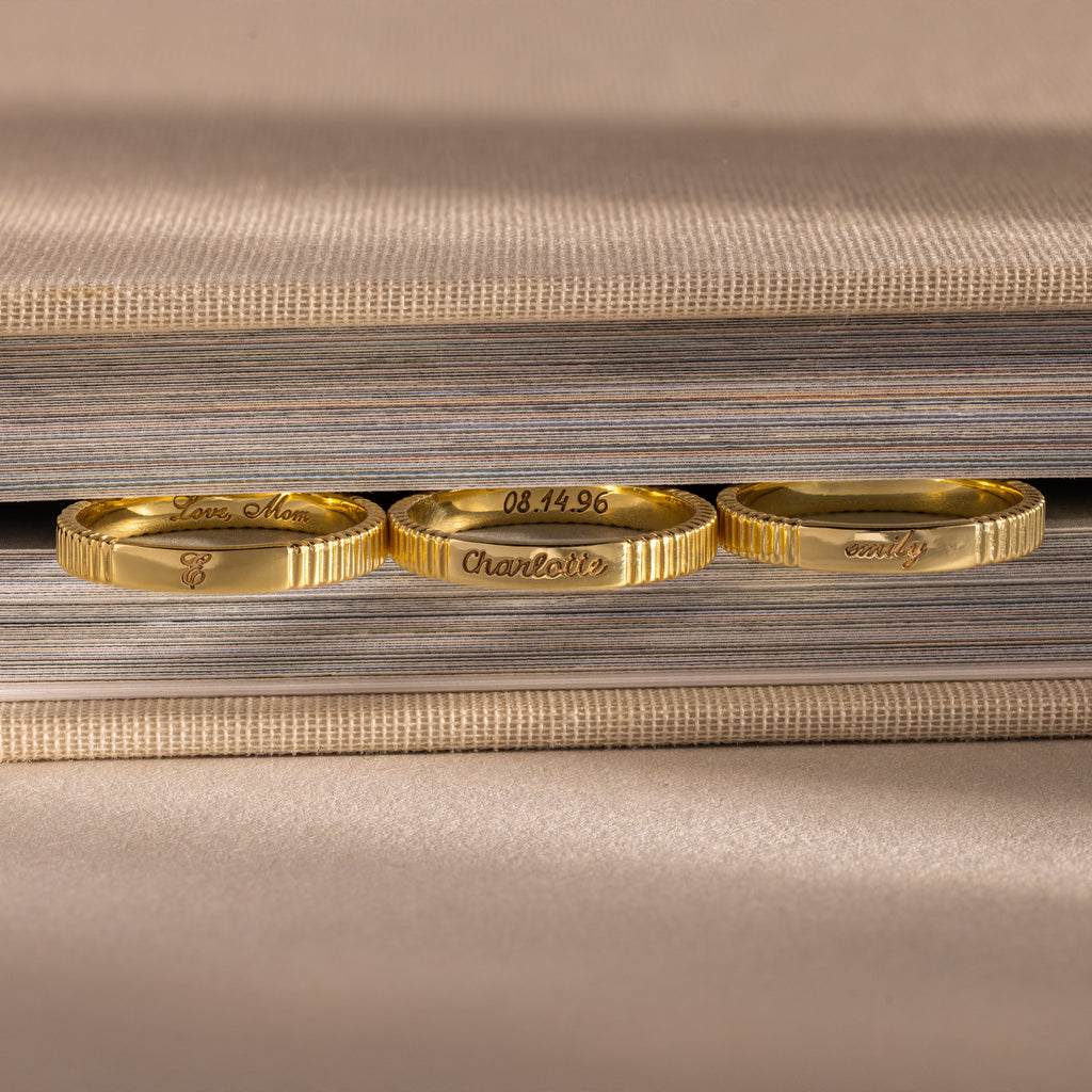 Three gold rings with engraved messages—including a custom piece and the Ribbed Name Ring in 18K Gold—rest upright against the edge of a closed book.