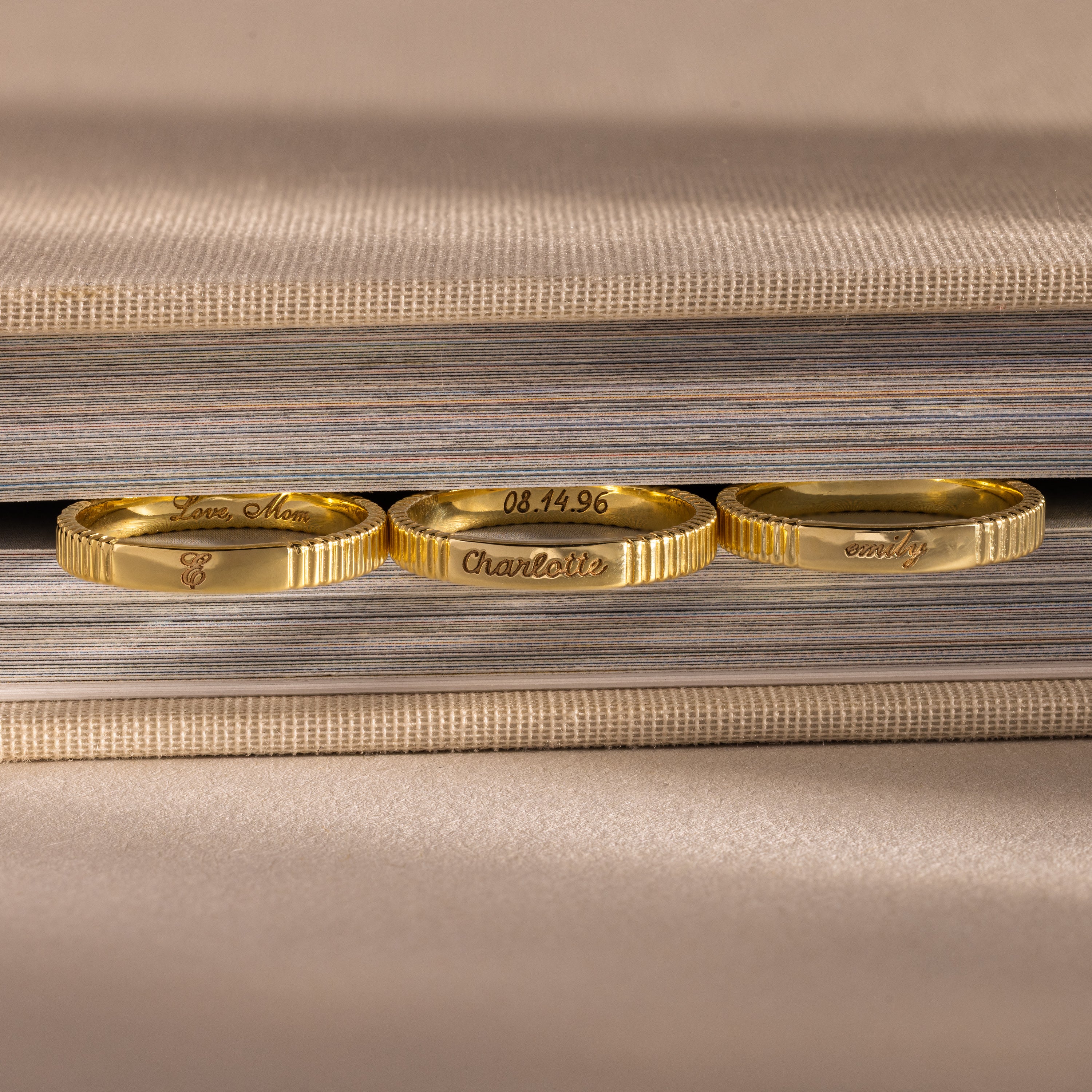 Three Ribbed Name Rings, each engraved with a custom name, rest on the edge of a closed, textured book.