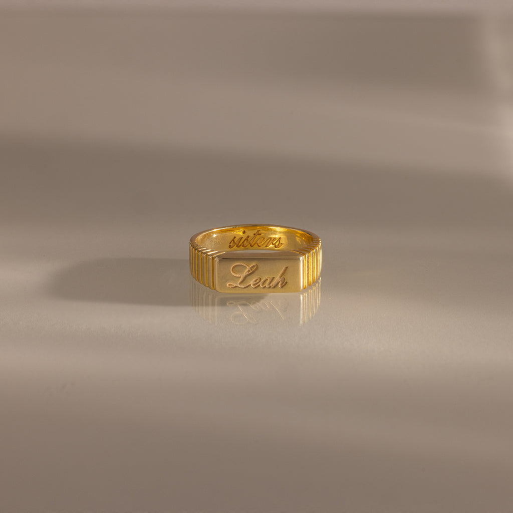 A Ribbed Name Signet Ring engraved with “victor” and “Leah” rests on a smooth, light surface.