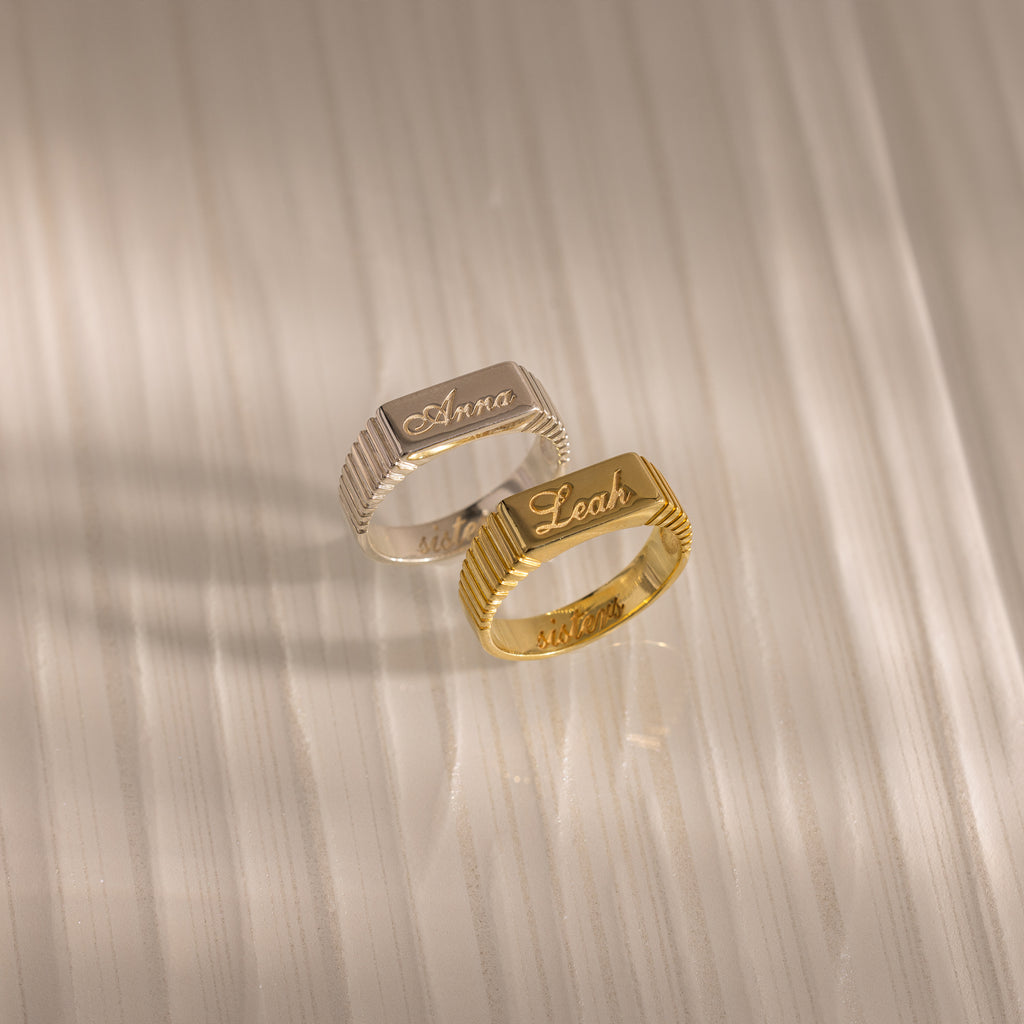 Two Ribbed Name Signet Rings, one silver engraved with "Anna" and one gold with "Leah," rest on a textured light surface—each personalized ring is a unique treasure.