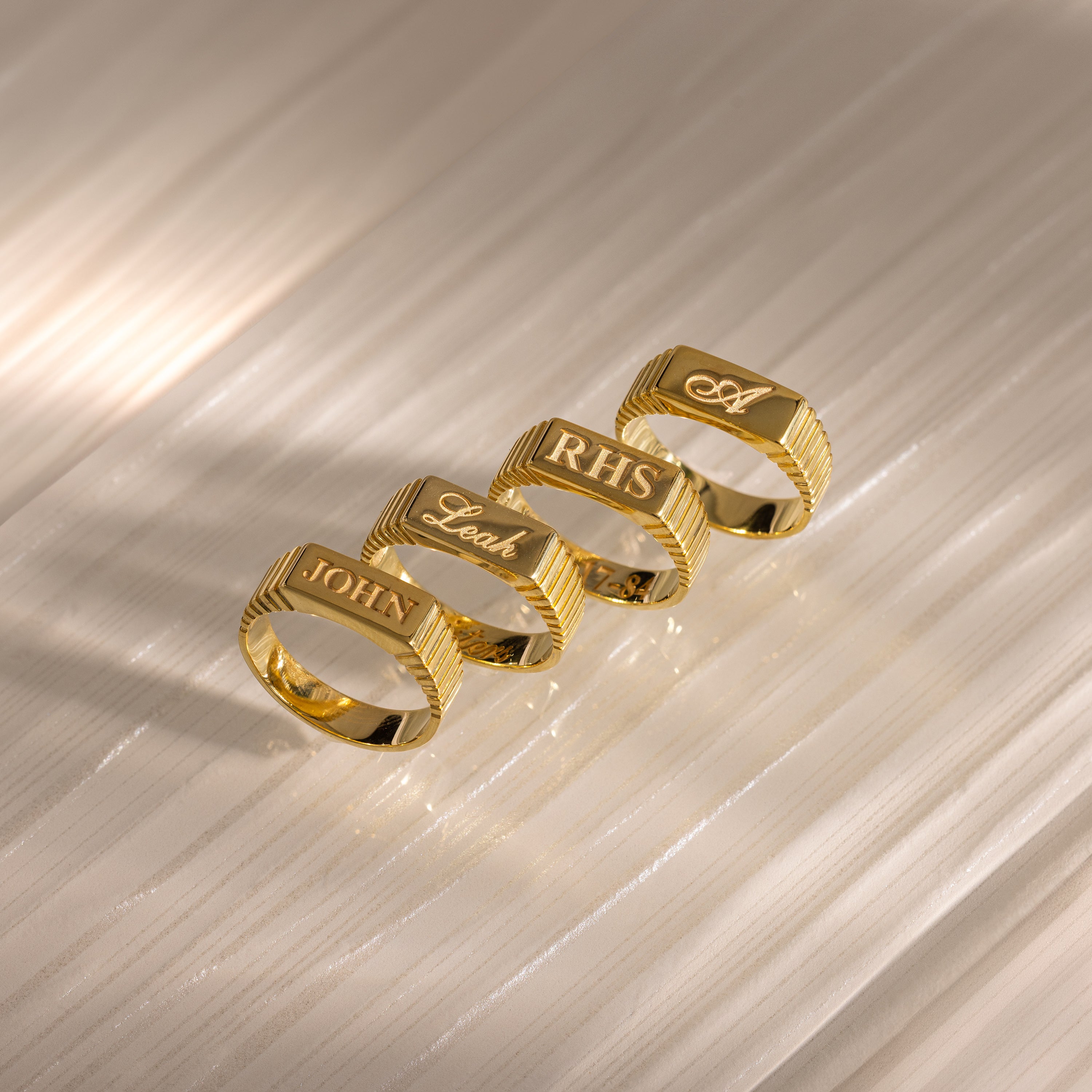 Four gold engraved signet rings, including the Ribbed Name Signet Ring, are arranged in a row on a light textured surface, each personalized with names or initials.