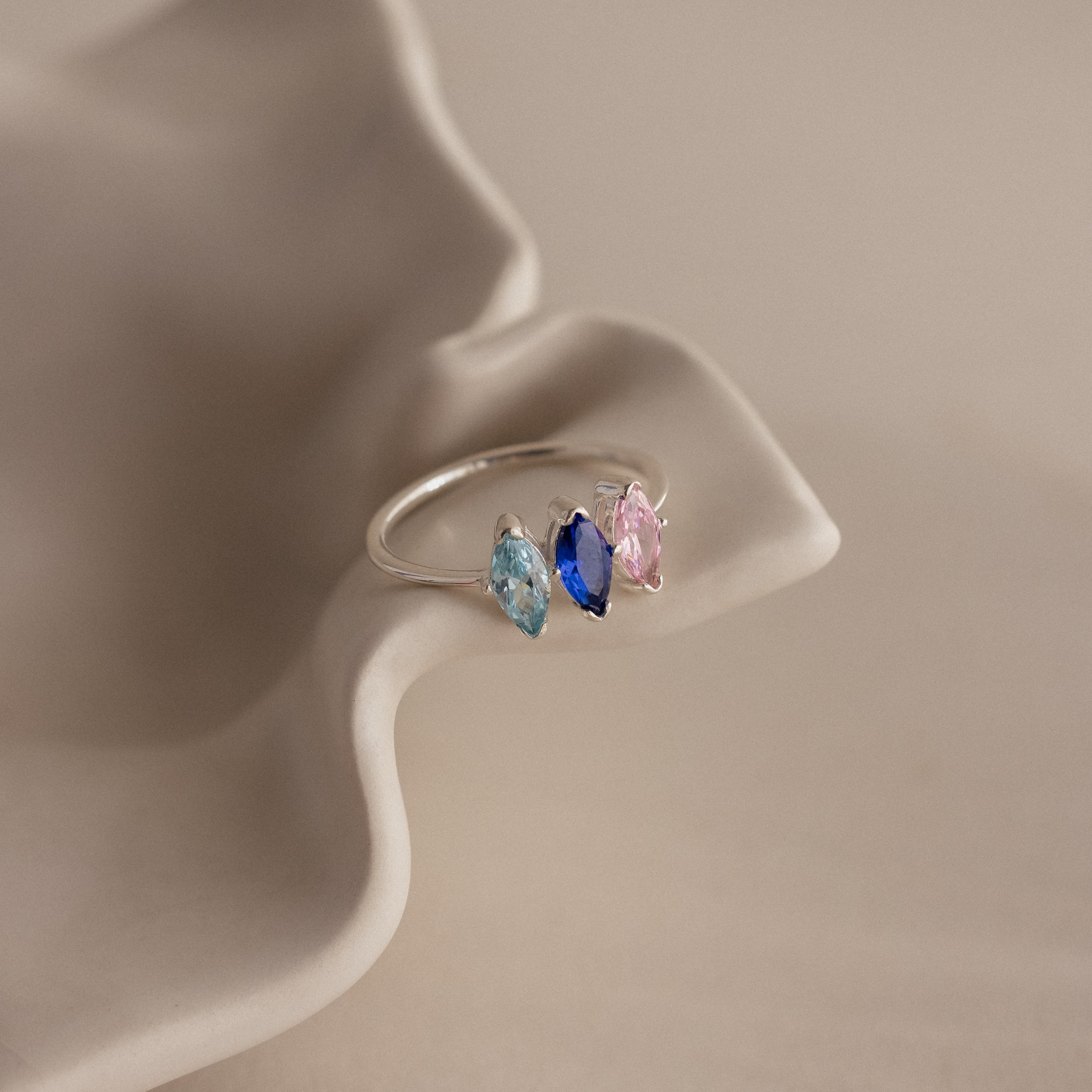 The Multiple Marquise Birthstone Ring in Sterling Silver features three marquise-cut stones—light blue, dark blue, and pink—on a wavy, beige surface. A personalized jewelry piece ideal for milestone gifts or special occasions.