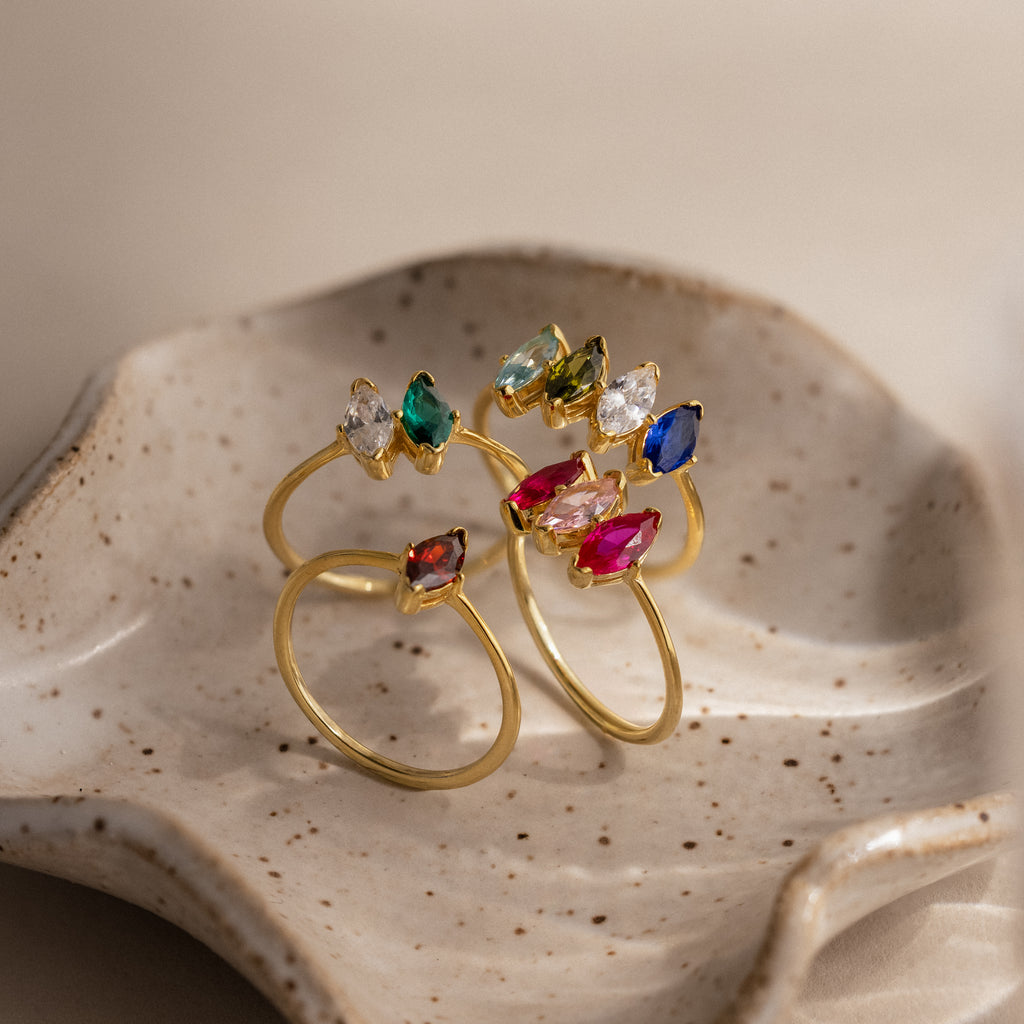 The Multiple Marquise Birthstone Ring, a gold ring featuring customizable marquise-cut stones, is displayed on a speckled ceramic dish.