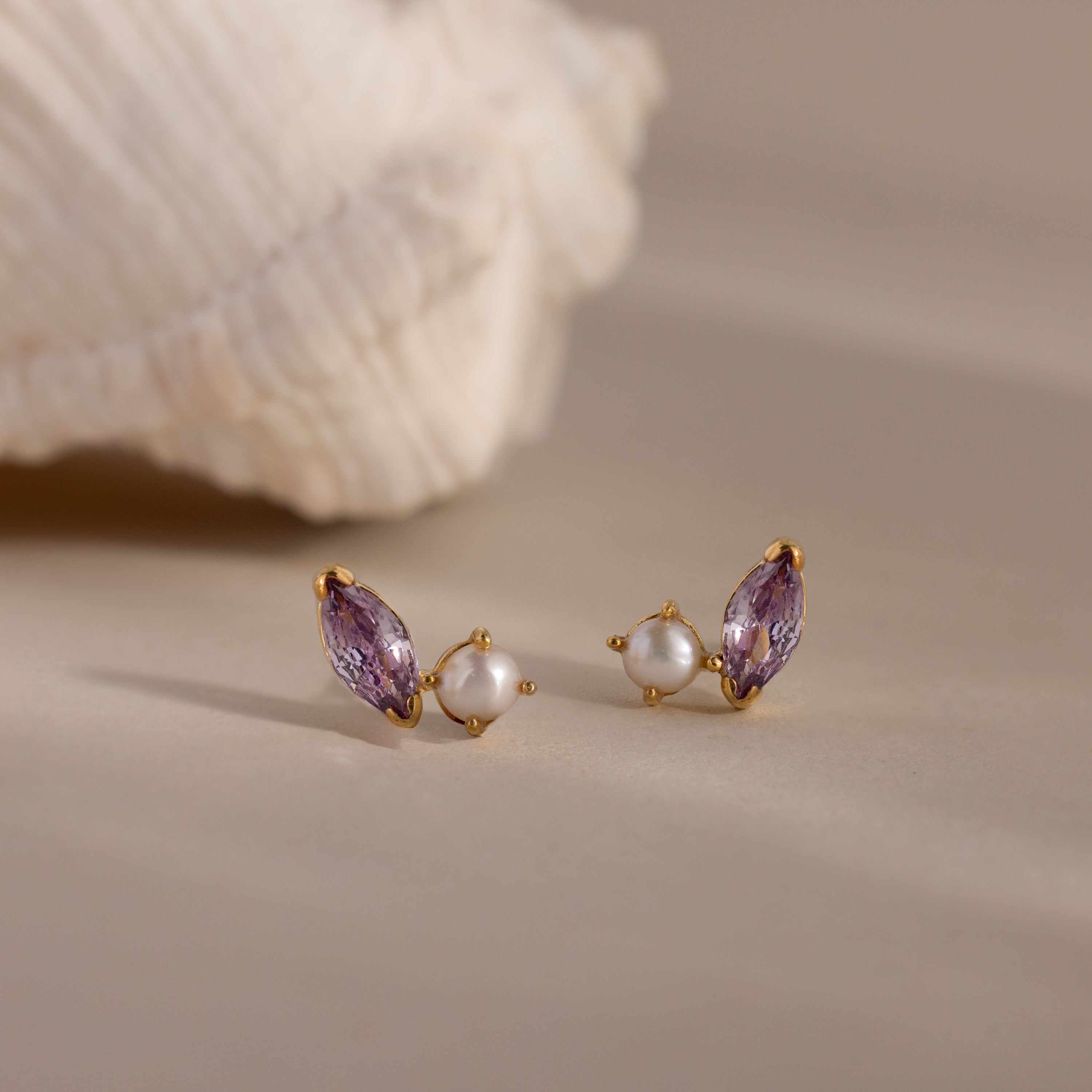 Marquise Alexandrite & Pearl Studs, featuring gold toi et moi design with purple gemstones and pearls, displayed on a neutral surface with a shell in the background.
