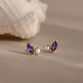 The Marquise Amethyst & Pearl Studs feature a rich purple marquise amethyst and delicate pearl accents, beautifully displayed on a neutral surface beside a shell.
