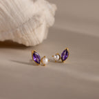 The Marquise Amethyst & Pearl Studs feature a rich purple marquise amethyst and delicate pearl accents, beautifully displayed on a neutral surface beside a shell.