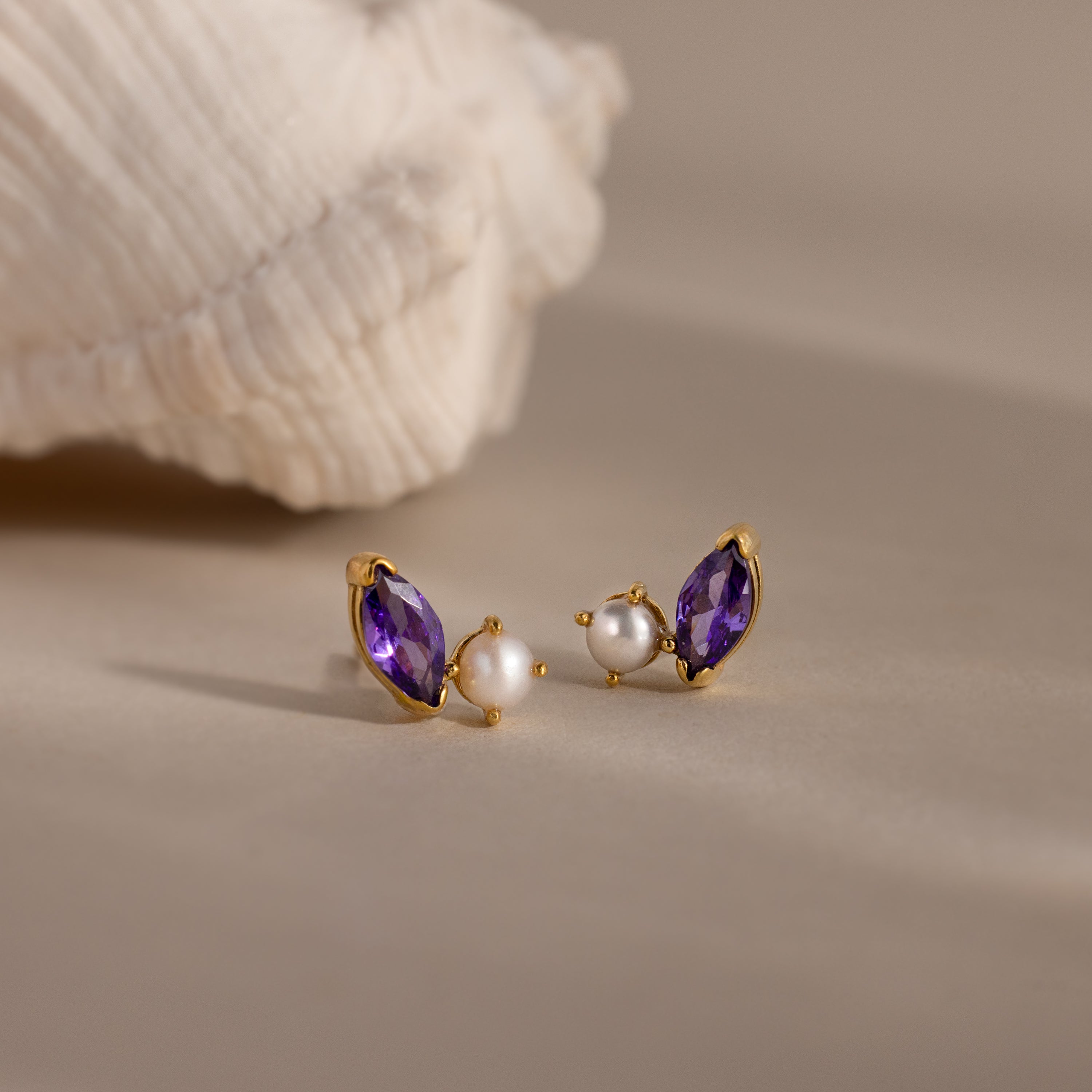 The Marquise Amethyst & Pearl Studs feature a rich purple marquise amethyst and delicate pearl accents, beautifully displayed on a neutral surface beside a shell.