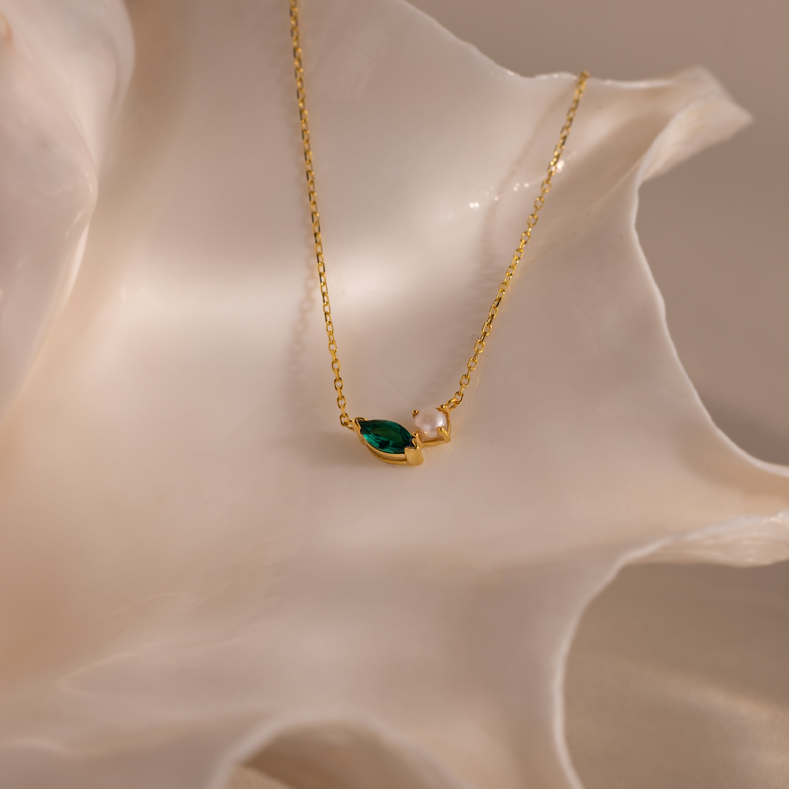 The Marquise Emerald & Pearl Necklace, featuring a green marquise birthstone and a small pearl, is displayed on a white seashell backdrop.