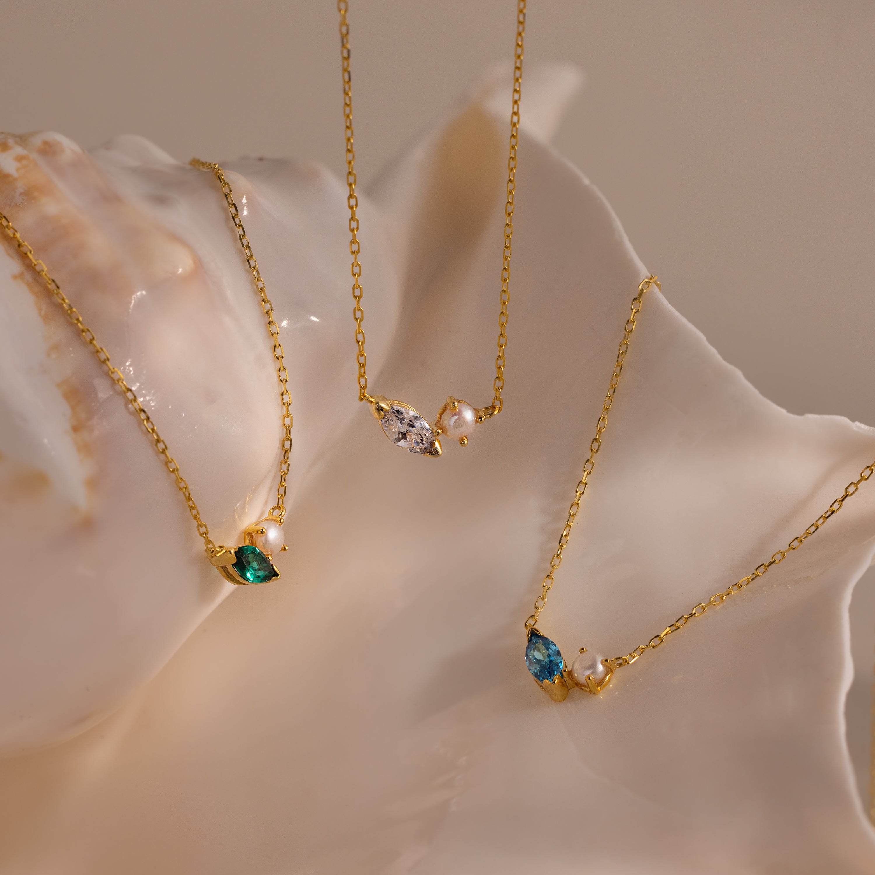The Marquise Birthstone & Pearl Necklace, along with two other gold jeweled pendant necklaces, are elegantly displayed on a large, glossy seashell.