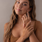 A woman with long hair smiles softly, touching her face as she wears a gold Marquise Birthstone & Pearl Necklace.