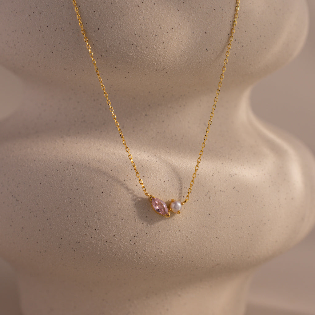 The Marquise Pink Tourmaline & Pearl Necklace features a gold chain with a pink marquise stone and small pearl pendant, displayed on textured beige. This elegant piece is a stunning customizable birthstone necklace for any occasion.